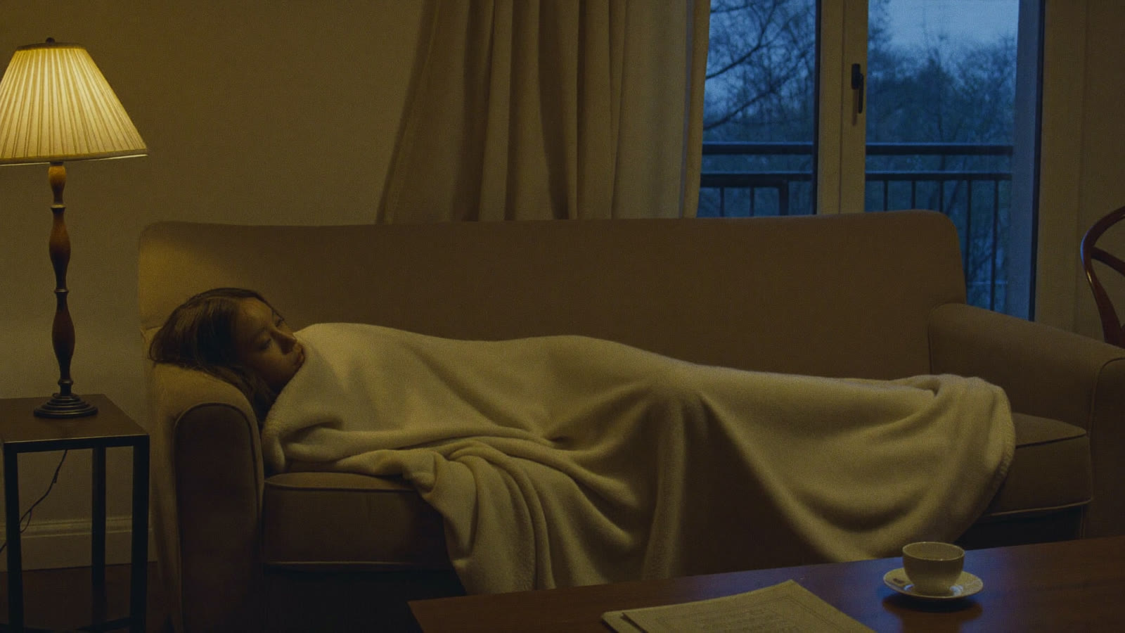 A person wrapped in a blanket on a couch between a warm lamp and cool window light